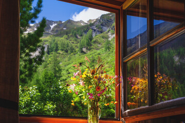Blick aus einer Almhütte in die Landschaft auf die Berge. Im Fenster steht ein bunter Wiesenstrauß