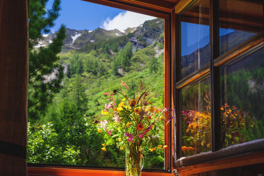 Blick aus einer Almh&uuml;tte in die Landschaft auf die Berge. Im Fenster steht ein bunter Wiesenstrau&szlig;