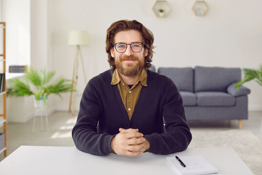 Portrait of happy smart businessman or teacher during video call or online conference. Handsome bearded man in eyeglasses and cardigan sitting at table in living room during videocall work meeting