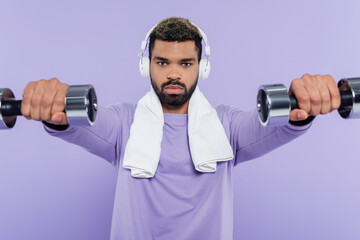 Fototapeta na wymiar young african american man in headphones exercising with dumbbells isolated on purple.