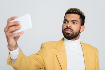 cheerful african american man in yellow blazer taking selfie on smartphone isolated on grey.