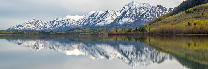 Panoramic view of a wilderness lake in Canada during spring with reflection of snow capped mountains in water. 
