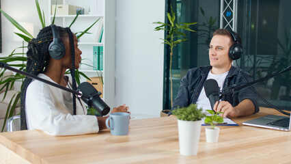 Fototapeta premium A Male Presenter Communicates with a Guest, an African American, During a Radio Broadcast at a Table in a Recording Studio, Broadcasts a Live Radio Interview With Spbd. Male Host Sharing Content.
