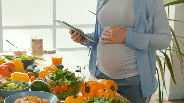 A Pregnant Woman Using a Tablet Prepares Healthy Food in a Bright Kitchen With a Modern Interior. Taking Care of Children. Woman Chooses the Right Food. Organic. Close-up.