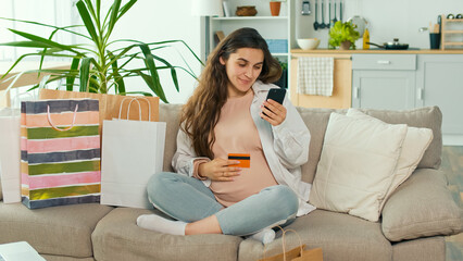 Young Pregnant Woman Makes Online Purchases Using her Phone and Credit Card in a Bright Living Room at Home with Designer Interiors. Online Delivery and Comfort Ordering Concept.