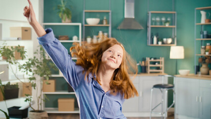 Cute Red-haired Teenage Girl Dancing Vigorously in Modern Kitchen at Home, Having Fun, Feeling Happy. Confident Teenager. Joyful Young People And Entertainment Concept. Mental Health.