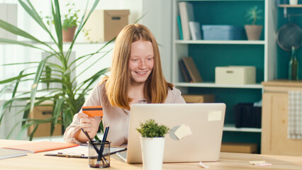 Happy Red-haired Teen Girl Shopping Online with a Card From Home. Consumer, Easy Shopping, Discounts, Online Shopping. Earlier Financial Development.