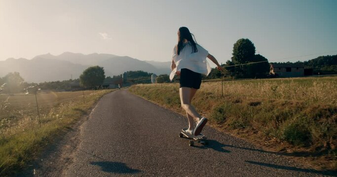Hipster girl ride on skate longboard on beautiful scenery at sunset along the meadow and mountain at background. Cinematic youth shot. Authentic candid portrait of millennial skateboarder woman