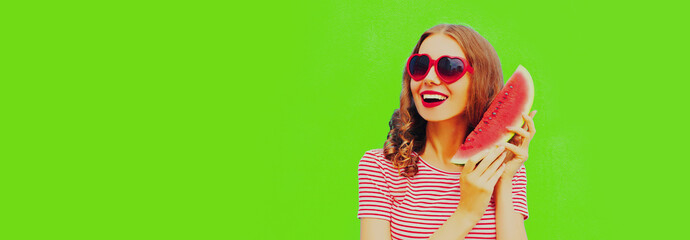 Portrait of happy smiling young woman with slice of watermelon wearing red heart shaped sunglasses on green background, blank copy space for advertising text