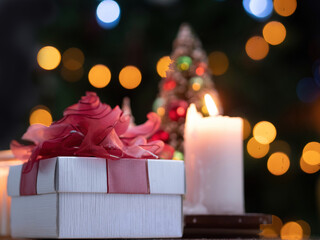 Selective focus, white gift box with red ribbon next to burning candle with blurry background of Christmas tree with bright bokeh.