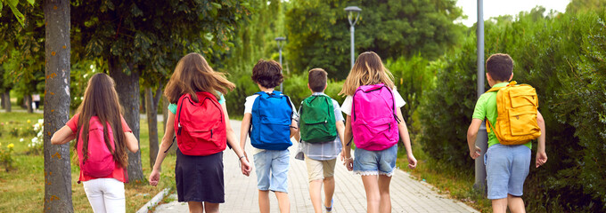 Fototapeta Children on their way to school. Group of little kids going to class. Six male and female students with colorful red, green, yellow, blue and pink backpacks walking along the park path. Banner, header obraz