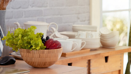 Selective focus, wooden basket filled with green lettuce leaves, capsicum with blurry background of white porcelain plates bowl and cup on kitchen counter.