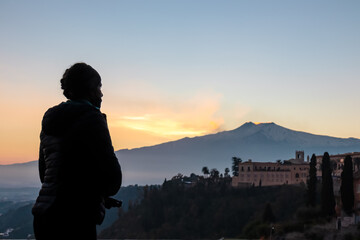 Fototapeta premium Tourist woman at a decorative railing with scenic view on snow capped Mount Etna volcano during sunset from public garden Parco Duca di Cesaro to Giardini Naxos in Taormina, Sicily, Italy, Europe, EU