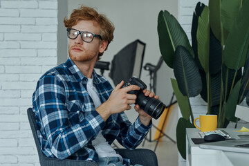 Confident young man holding digital camera while sitting at his working place in studio