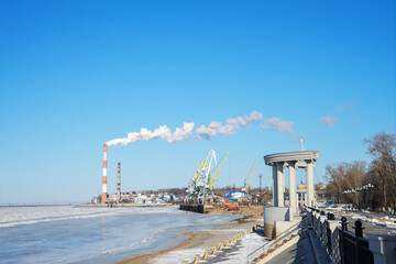 Obraz premium Embankment of Amur River, covered with ice on clear winter day, Khabarovsk. White smoke from chimneys of factories on riverbank, port cranes. Сoncept of ecology