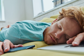 Tired redhead man napping on the couch with laptop and smart phone laying near him