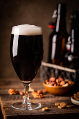 Glass of dark beer with foam head on dark wooden background with empty bottles and beer snacks