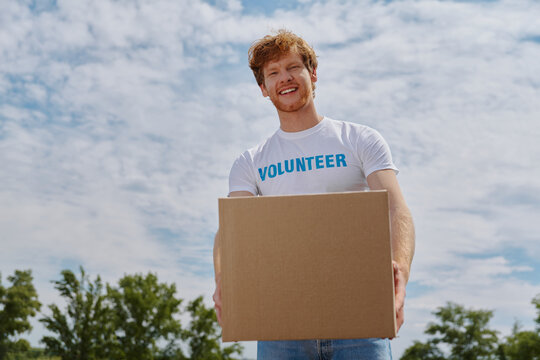 Handsome young man in volunteer shirt carrying box and smiling while standing outdoors