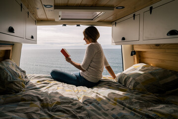 Concept: Camper van. Girl reading a book in the bed of the motorhome with open doors in seascape.