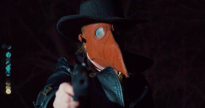Person in a hat, leather coat and a plague doctor mask aims a gun at a victim or enemy, and then shoots, front view. Criminal hid face so that he would not be recognized during robbery