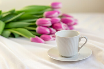 Fototapeta na wymiar A white cup with a saucer and a bouquet of beautiful pink tulips lies on a white tablecloth.
