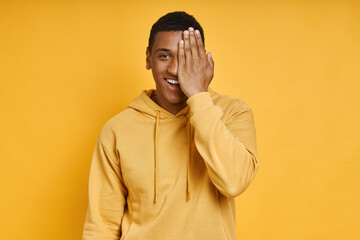 Playful mixed race man covering half face with hand while standing against yellow background