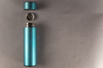 A turquoise metal thermos wit lid and teapot lies against the gray background. A cylindrical vacuum flask. Container for hot and cold drinks. Equipment for sports, travel, hiking and tourism.