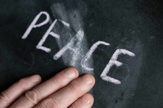 The semi-erased word PEACE on the black chalkboard. An adult man's left hand removes the handwritten word with his fingers. Fuzzy letters on black surface.