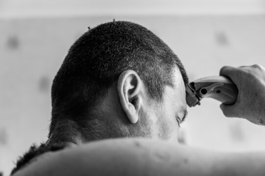 A middle-aged man cuts his hair. The person cuts the hair on his head with an electric hair clipper. Short brown hair. Inside a living room. Selective focus. Black and white photo.