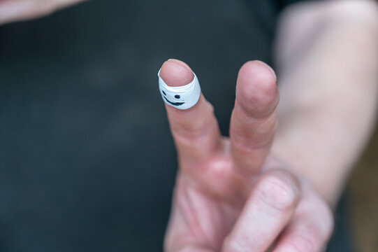 Close-up Of A Man's Hand Showing A Gesture Of Victory. White Armband On Adult Male Index Finger. Smiling Face Painted On The Armband. Positive Emotion Concept. Indoor. Selective Focus.