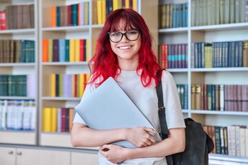 Portrait of teenage student female with backpack laptop looking at camera in college library