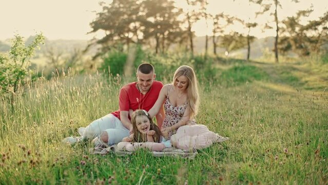 Young mom and dad with cute little daughter having fun outdoors in the woods. Little daughter in a white summer dress lies on the grass and laughs happily, mother tickles her daughter