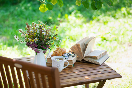 Bouquet of meadow flowers, croissant, cup of tea or coffee, books on table in summer garden. Rest in garden, reading books, breakfast, vacations in nature concept. Summertime in garden on backyard