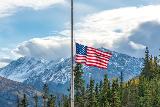 United States of America flag flying half mast with blue sky, clouds and mountain background in the north during fall, autumn season.