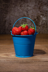 Ripe red strawberries in a toy blue bucket on a wooden table against a dark background. Close up.