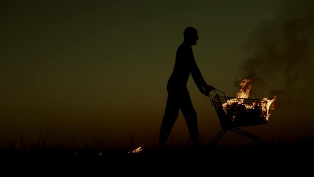 A man rolls a burning cart from a supermarket in an empty field, against the background of a golden sunset closer to the dark night, slow motion