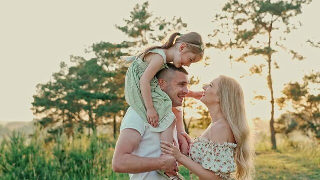 Young married couple with cute girl spend time together outdoors . The father holds daughter around his neck, the mother stands nearby and smiles, the daughter gently strokes her mother's face