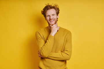 Cheerful redhead man holding hand on chin while standing against yellow background