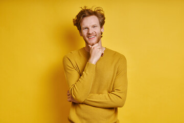 Fototapeta premium Cheerful redhead man holding hand on chin while standing against yellow background