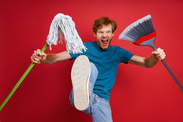 Furious young man carrying cleaning equipment and kicking while against red background