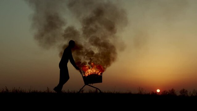 A man rolls a burning cart from a supermarket in an empty field, against the background of a golden sunset closer to the dark night, slow motion