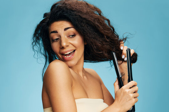 Beautiful smiling attractive Latin curly woman using hair straightener, looking aside, posing isolated on blue wall background. Hair routine concept, haircare, dry damaged hair ironing, hairdressing