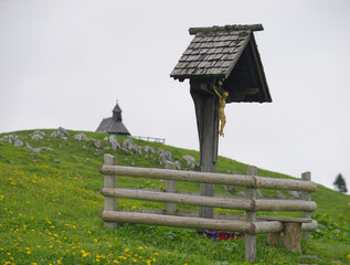 Fototapeta na wymiar Wooden cross in the mist and the church in the background, Velika Planina, Slovenia