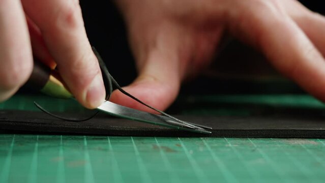 Man tailor cutting artificial leather close-up. Craftsman carving black belt at workplace table, making hand made products made of genuine animal leather. Professional atelier.