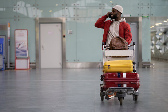 Young smiling African American man pushing luggage trolley while walking after arrival at airport, talking on mobile phone. Happy Black male tourist rolling a baggage cart in terminal. Trip, journey.