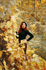 Stylish model among yellow dry foliage of trees. Beautiful female in black beret, knitted sweater and leather skirt. Awesome red-haired woman in autumn park. Walk in fall nature.