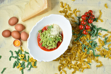 Fusilli dish in tomato sauce in a white plate against the background of flour, eggs, tomatoes, cheese, pasta