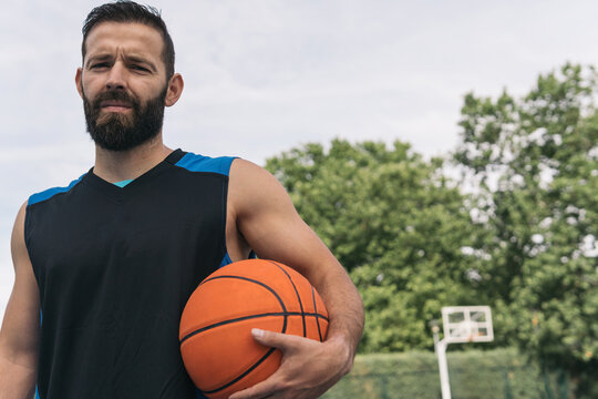 Young guy with a beard and a basketball under his arm looking at the camera seriously. Frontal close-up image of a man with a sleeveless shirt with an orange ball waiting for a street basketball game