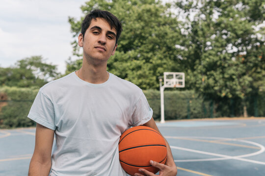 Young dark-haired boy with a basketball under his arm looking at the camera defiantly. Frontal close-up image of a man in a light-colored t-shirt posing with an orange ball waiting his rival