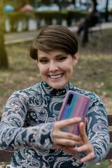 Caucasian woman with piercing and brown hair in a park taking a selfie.